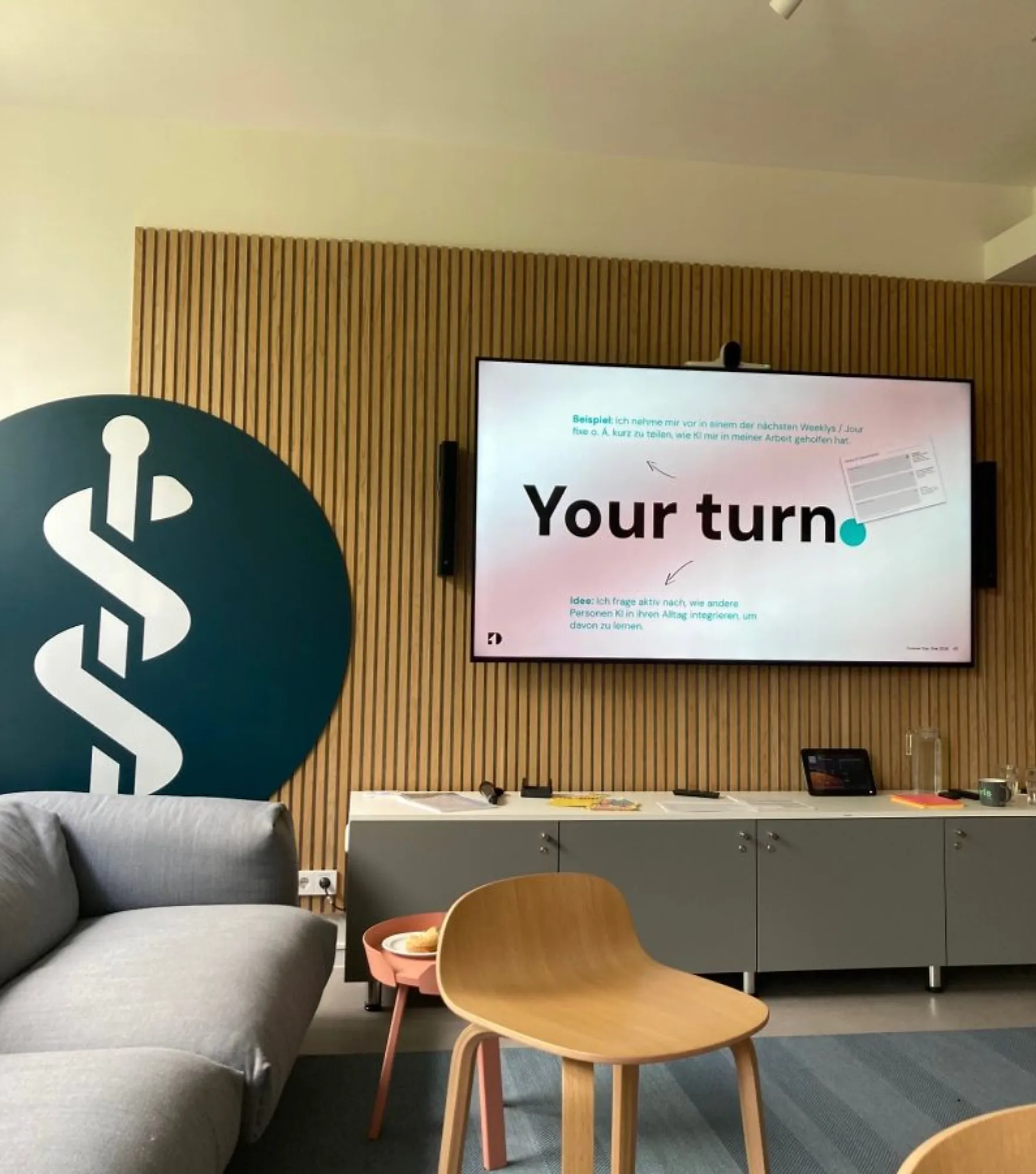 Workshop-Raum bei Asklepios: Auf dem Bildschirm der Impuls „Your turn.“, daneben das Asklepios-Logo an der Holzwand