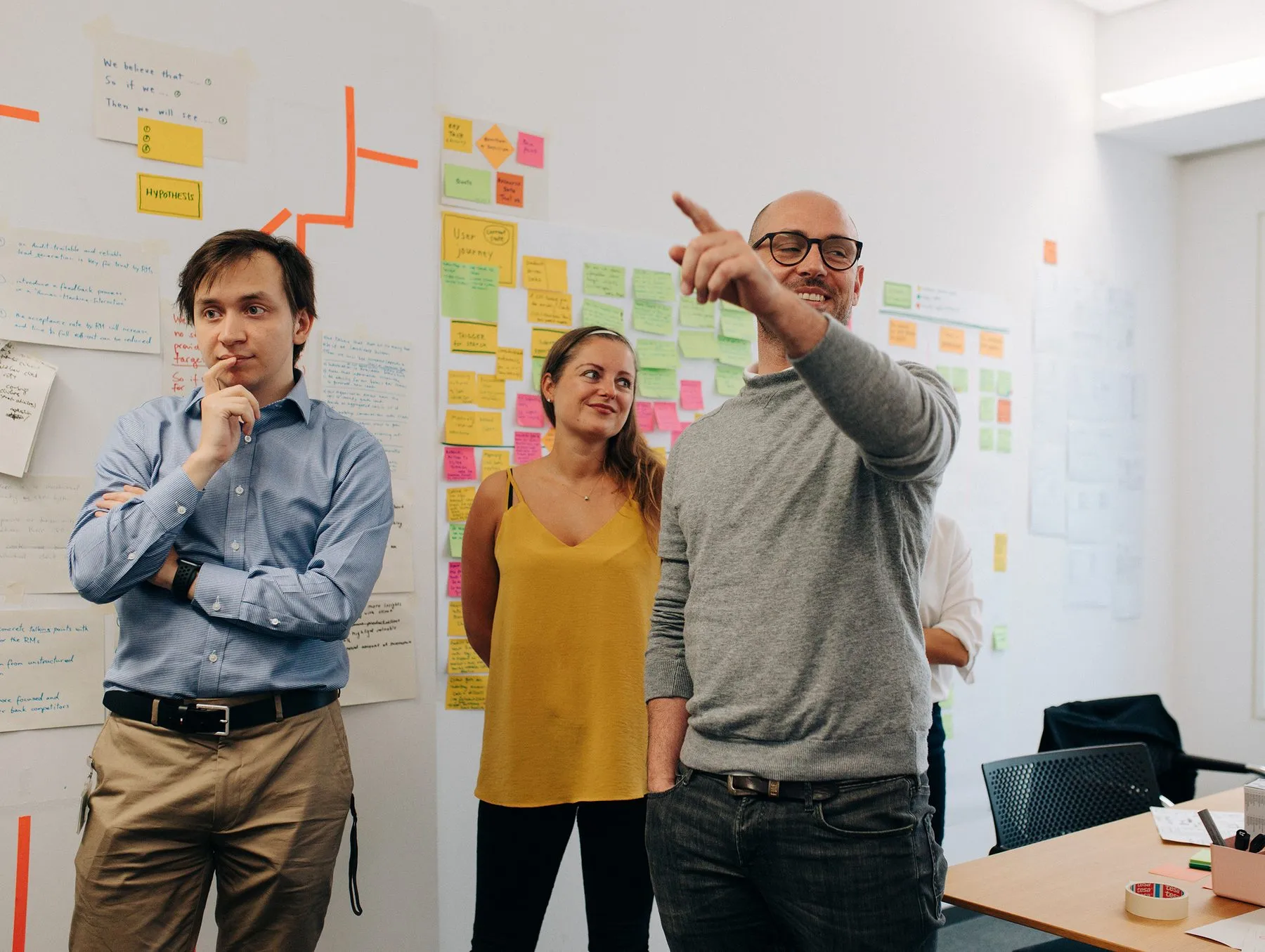 Forever Day One im Innovation-Space der Deutschen Bank: Ein Facilitator zeigt auf eine Wand voller Hypothesen und Post-its, zwei Teilnehmende hören zu