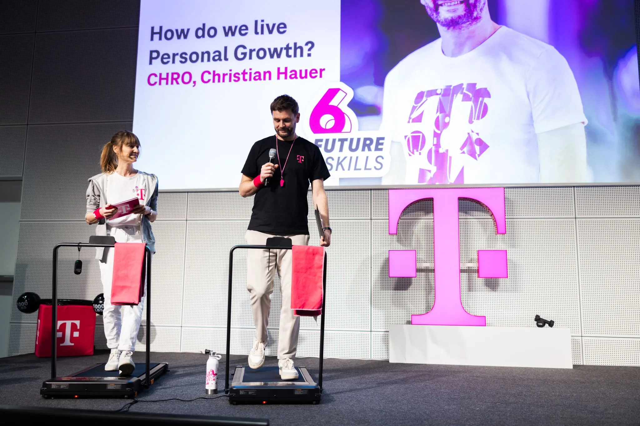 Zwei Personen auf Laufbändern auf der Bühne einer Future-Skills-Power-Gym-Session, im Hintergrund eine Leinwand mit „6 Future Skills“-Branding und ein großes pinkes T als Bühnenbild
