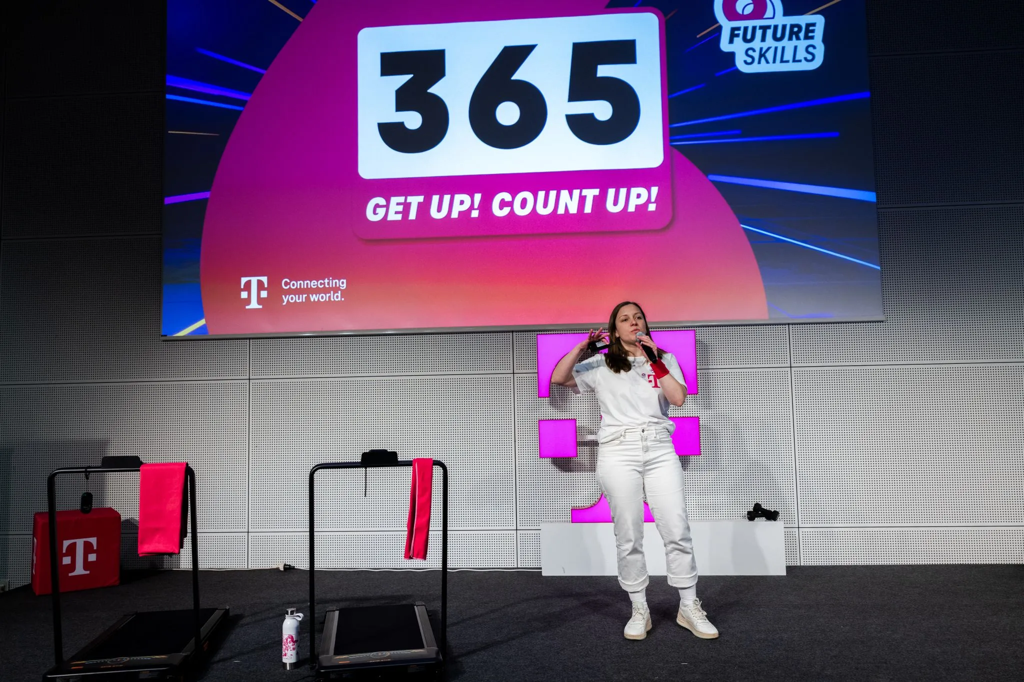 Weite Bühnenansicht einer Future-Skills-Power-Gym-Session: eine Sprecherin mit Mikrofon vor pinkem Bühnenbild, im Hintergrund eine Leinwand mit „365 – Get up! Count up!“ und dem Future-Skills-Logo